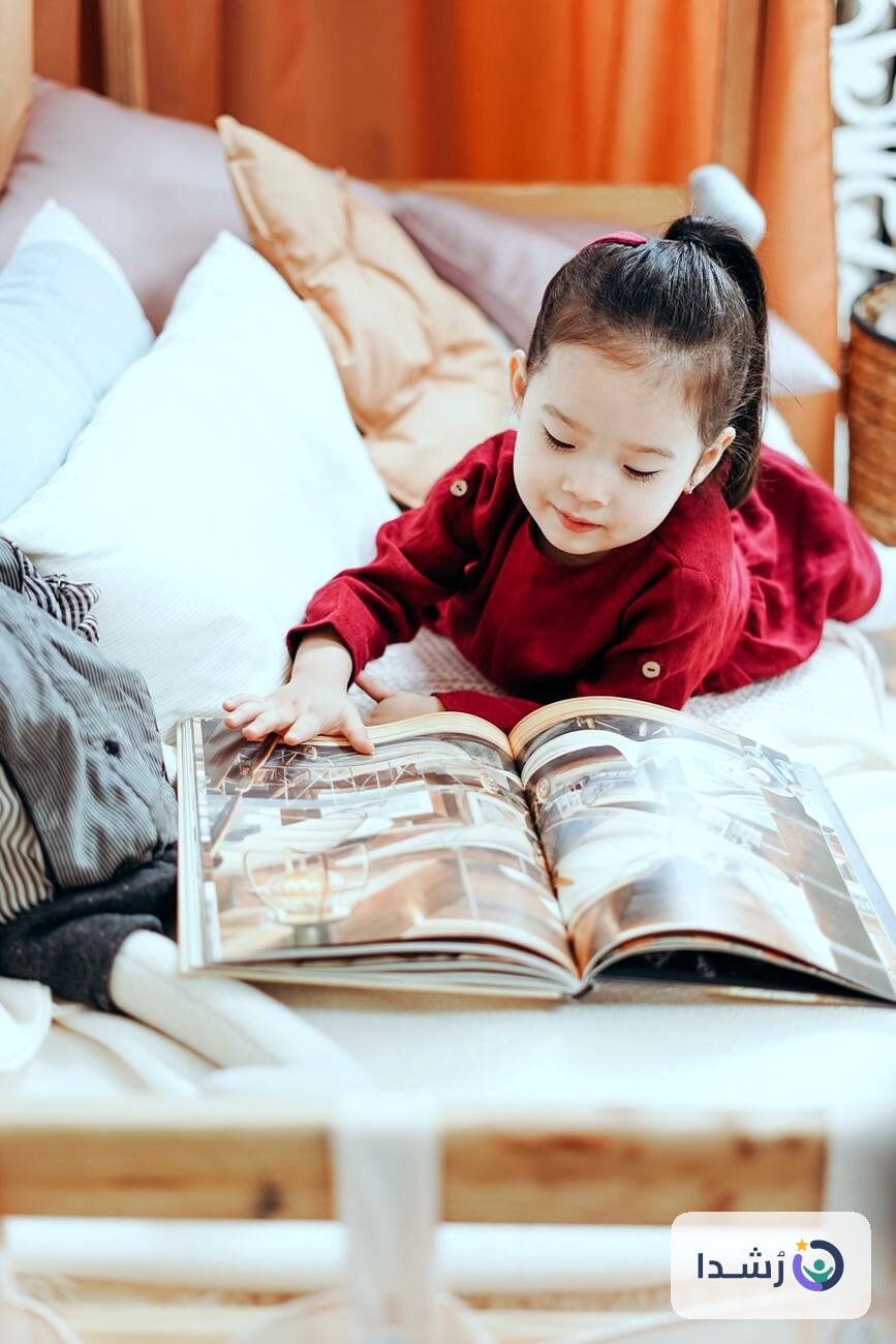 طفلة صغيرة تقرأ كتابًا ملونًا في غرفة مريحة.