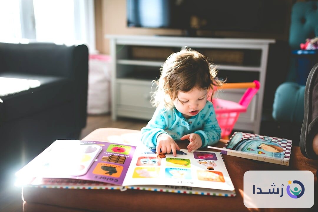 طفلة صغيرة تتعلم من كتاب ملون في غرفة معيشة مريحة.