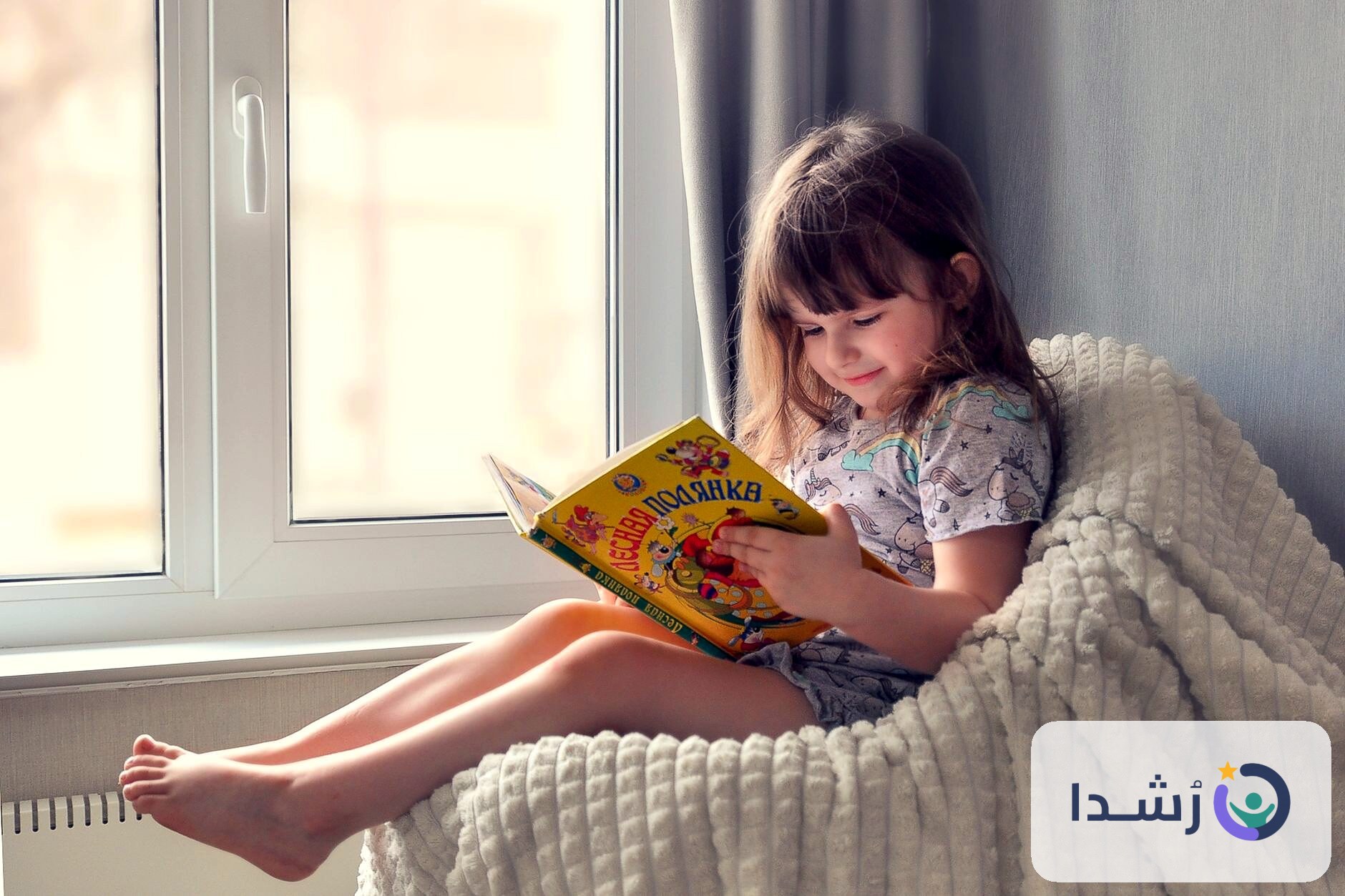 طفلة صغيرة تقرأ كتابًا ملونًا بجوار نافذة