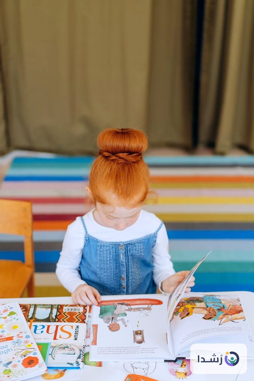 طفلة تقرأ كتابًا ملونًا على طاولة في غرفة مضاءة جيدًا.