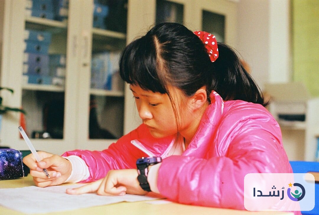 طفلة ترتدي سترة وردية وتكتب بتركيز في فصل دراسي.