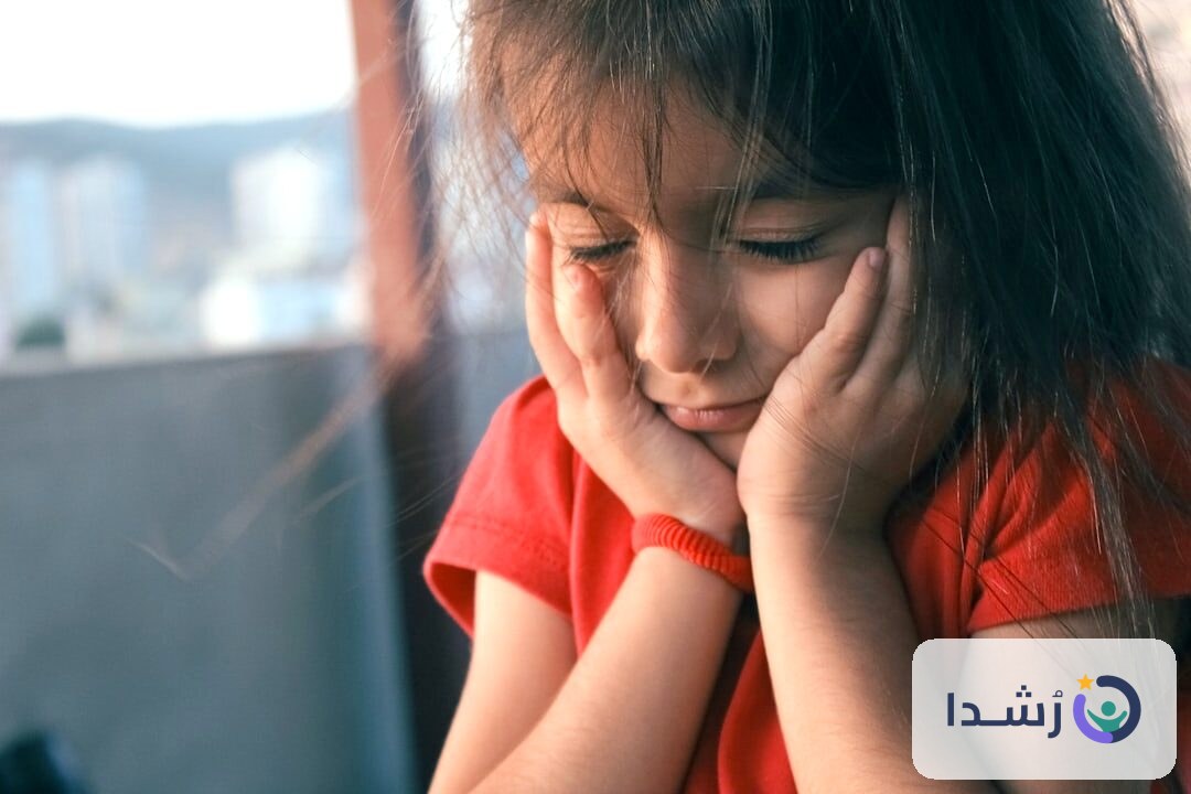 طفلة صغيرة تبدو حزينة وهي تمسك وجهها بيديها.